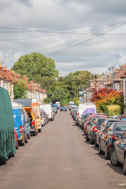 A residential street scene showing a narrow street lined with closely packed parked vehicles including small vans, cars, and delivery trucks, some of which are covered with protective tarps or plastic